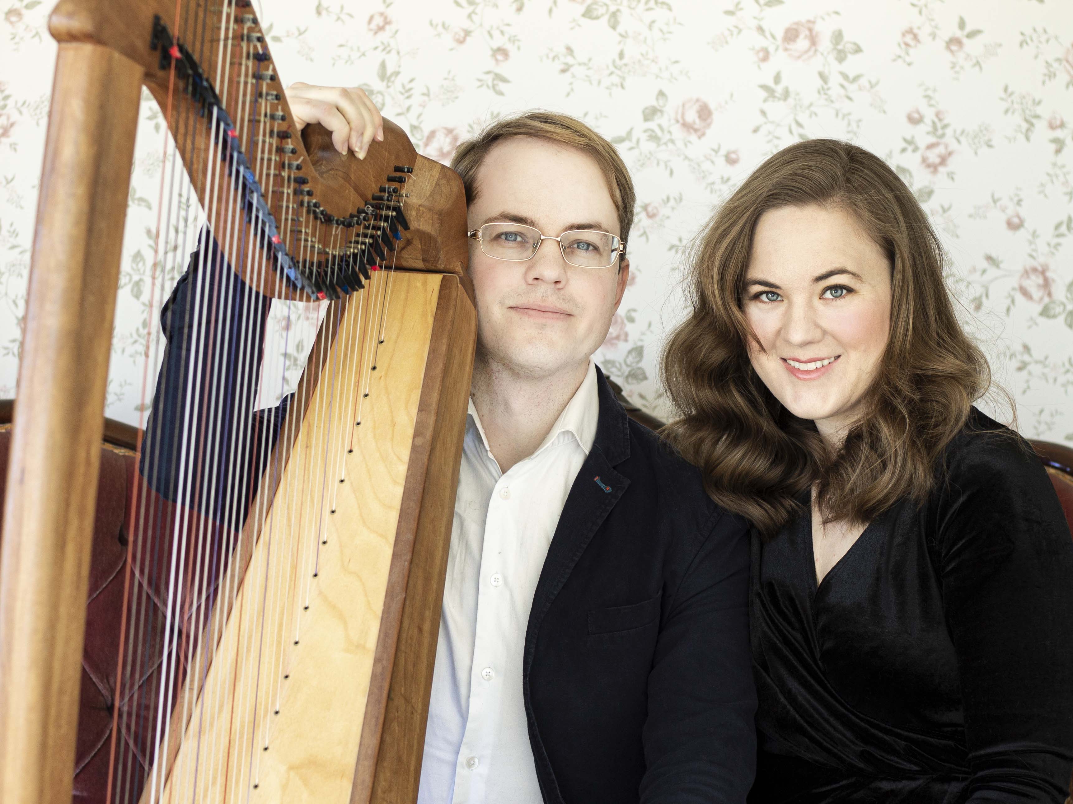 En man och en kvinna sitter i ett rum med blommig tapet och mannen håller vänster hand på en keltisk harpa.